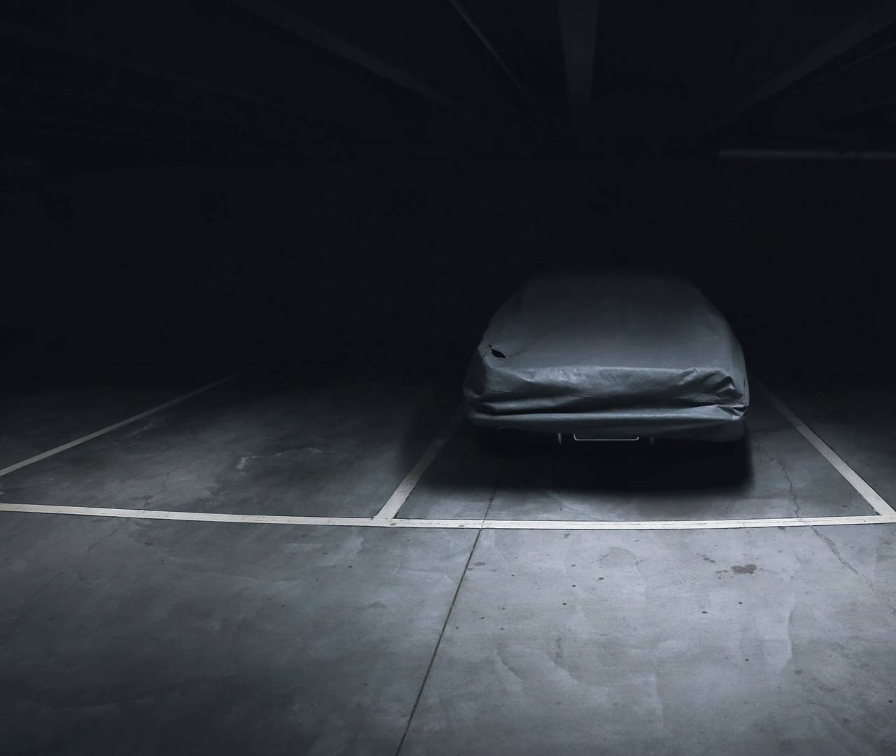 A covered car under a single light in an empty garage
